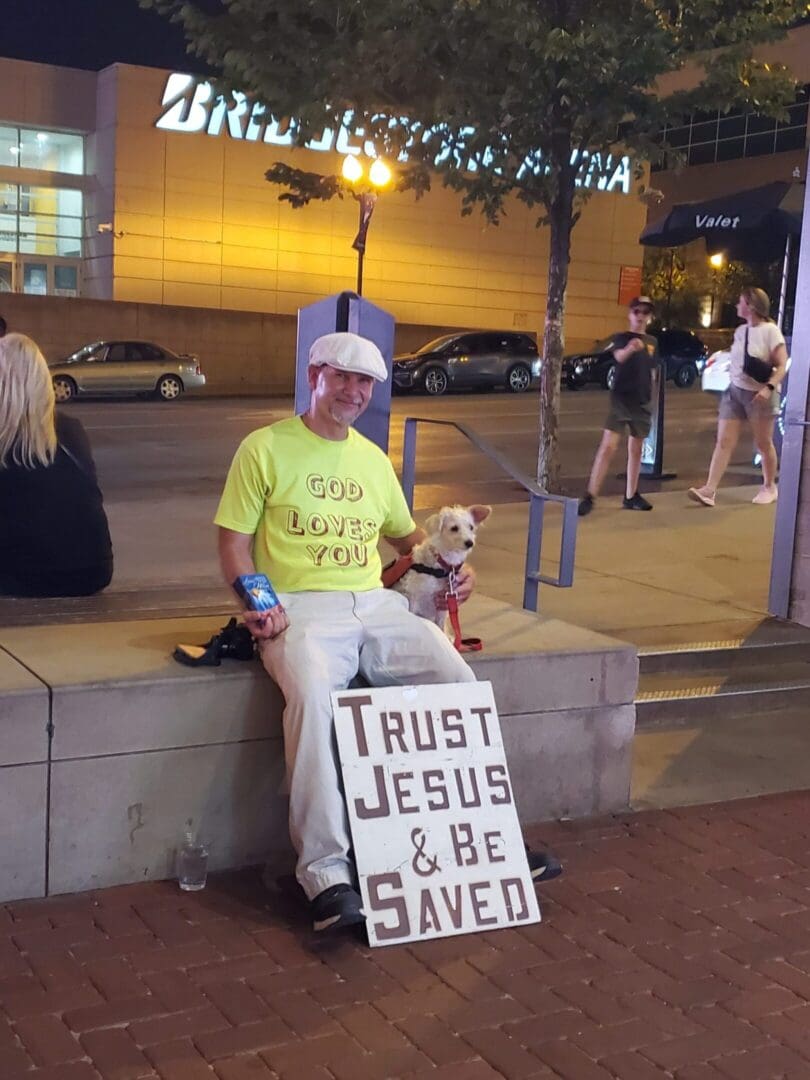 Man sitting with a sign that says 'Trust Jesus & Be Saved'.