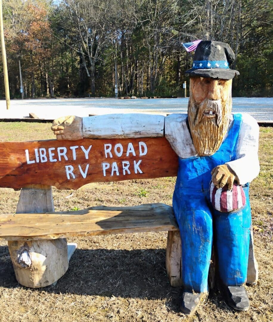 A wooden statue of a man with a beard sitting beside a Liberty Road RV Park sign.
